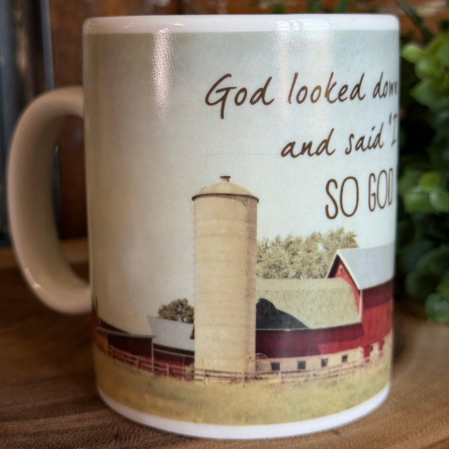 Rustic farmhouse, red barn, grain silo coffee mug with silo and red barn illustration and “God looked down… so God made a farmer” quote, displayed on a wooden surface with greenery in the background.