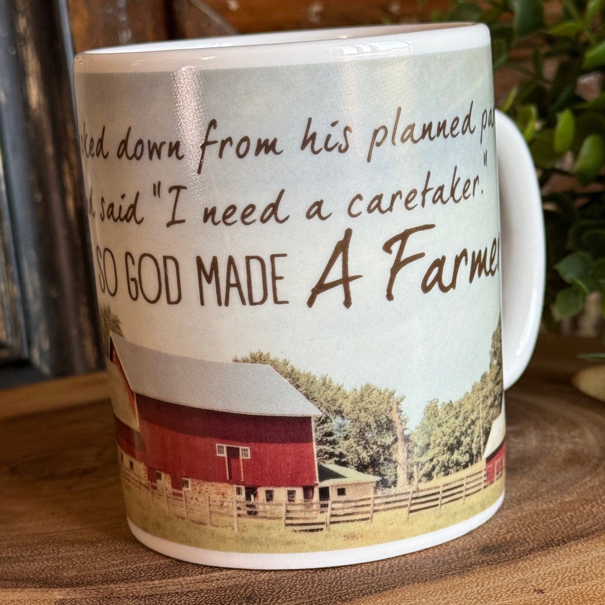 Rustic farmhouse coffee mug with red barn illustration and inspirational “God made a farmer” quote, sitting on a wooden surface with greenery in the background.