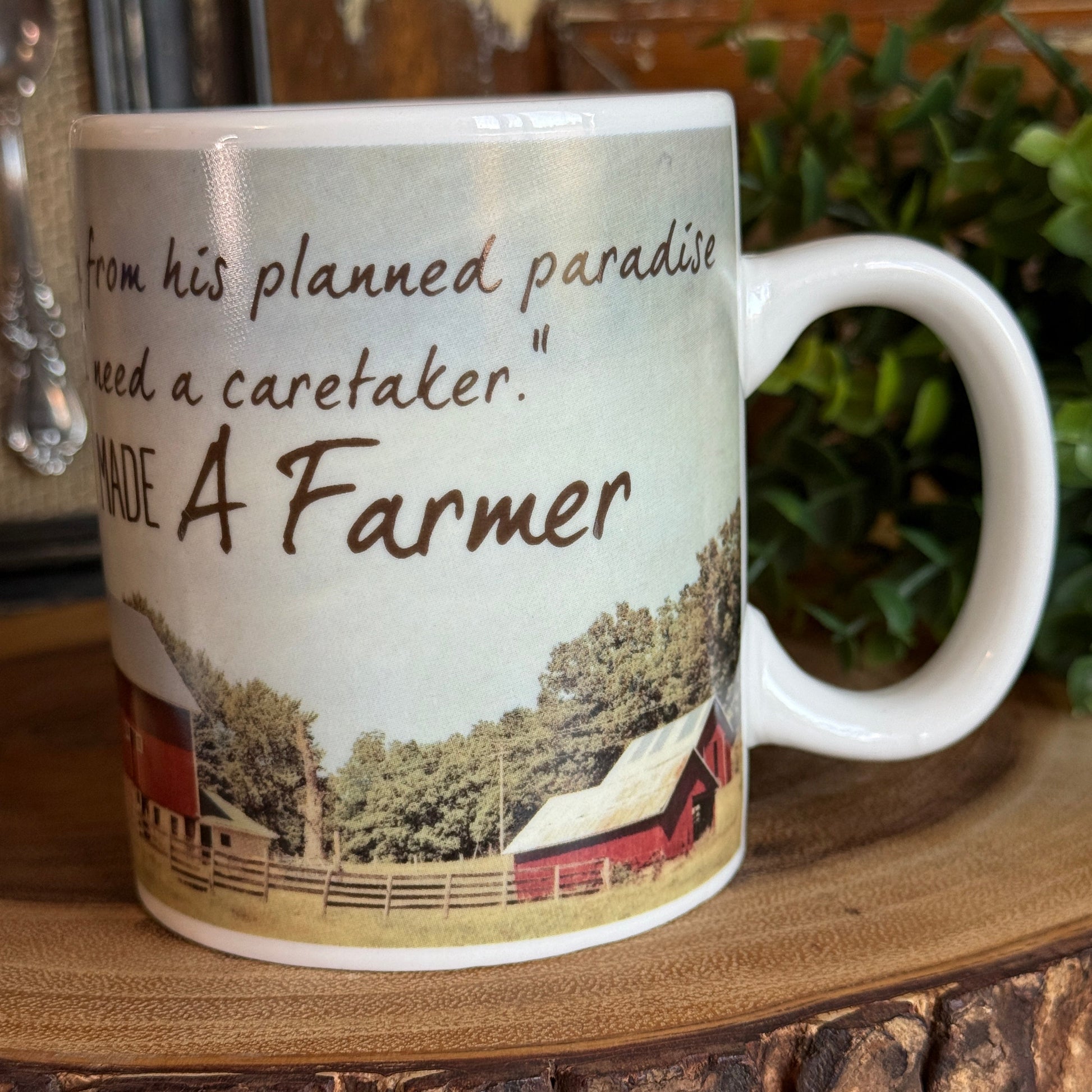 Rustic farmhouse coffee mug with red barn scene and “God made a farmer” quote, shown on a wooden slab surface with greenery in the background.